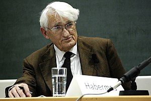 Picture of a man with white hair and glasses sitting behind a glass of water with a blackboard behind him.
