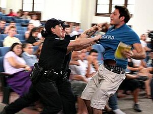 Two police officers in black uniforms grab the blue shirt of a man while onlookers sit around.